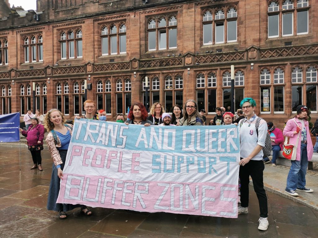 A number of LGBT+ people holding up a banner that reads "Trans and Queer people support buffer zones"