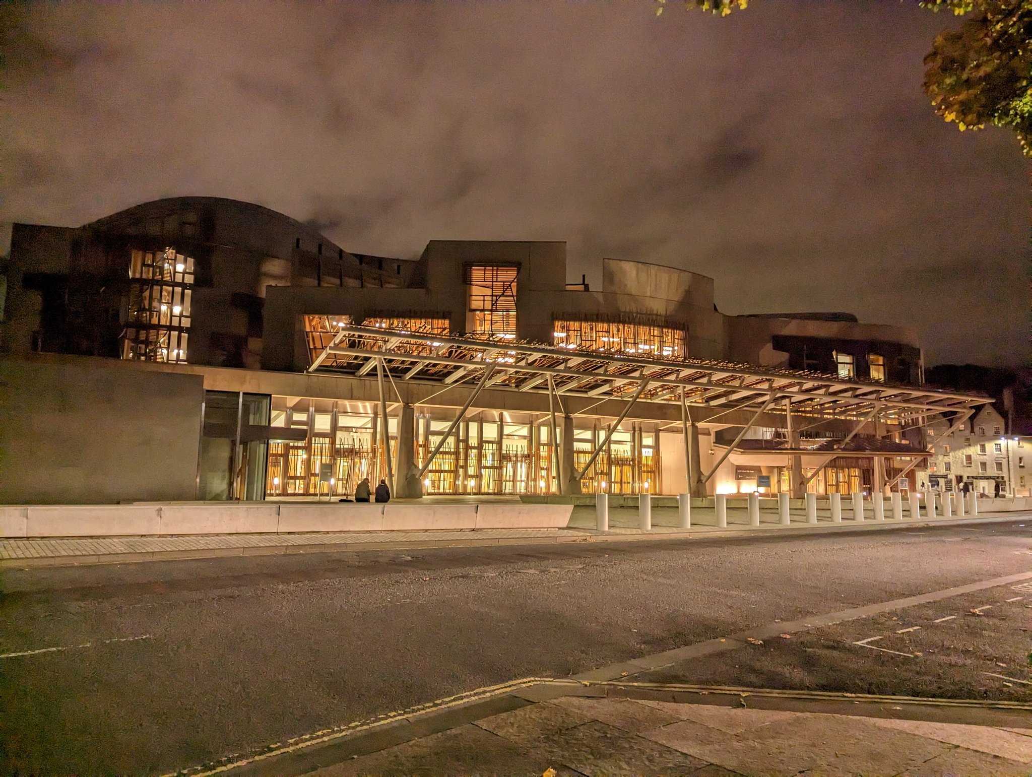 The image shows a large building, The Scottish Parliment, at nighttime with illuminated windows. The architecture features a mix of curved and angular elements, with some parts of the building having what appears to be metal framework visible from the outside. A canopy-like structure covers an area in front of the building, likely indicating an entrance or a covered walkway, with lights casting a warm glow underneath it. The sky is dusky or possibly overcast. In front of the building, there is a street with no visible traffic at the moment, and a pedestrian area with bollards demarcating the edge of the sidewalk. There also appears to be a lone person standing under the canopy structure. The overall scene is quite peaceful and deserted, which is common for urban areas during nighttime hours. The style of the building suggests it might be a modern cultural, educational, or business facility.