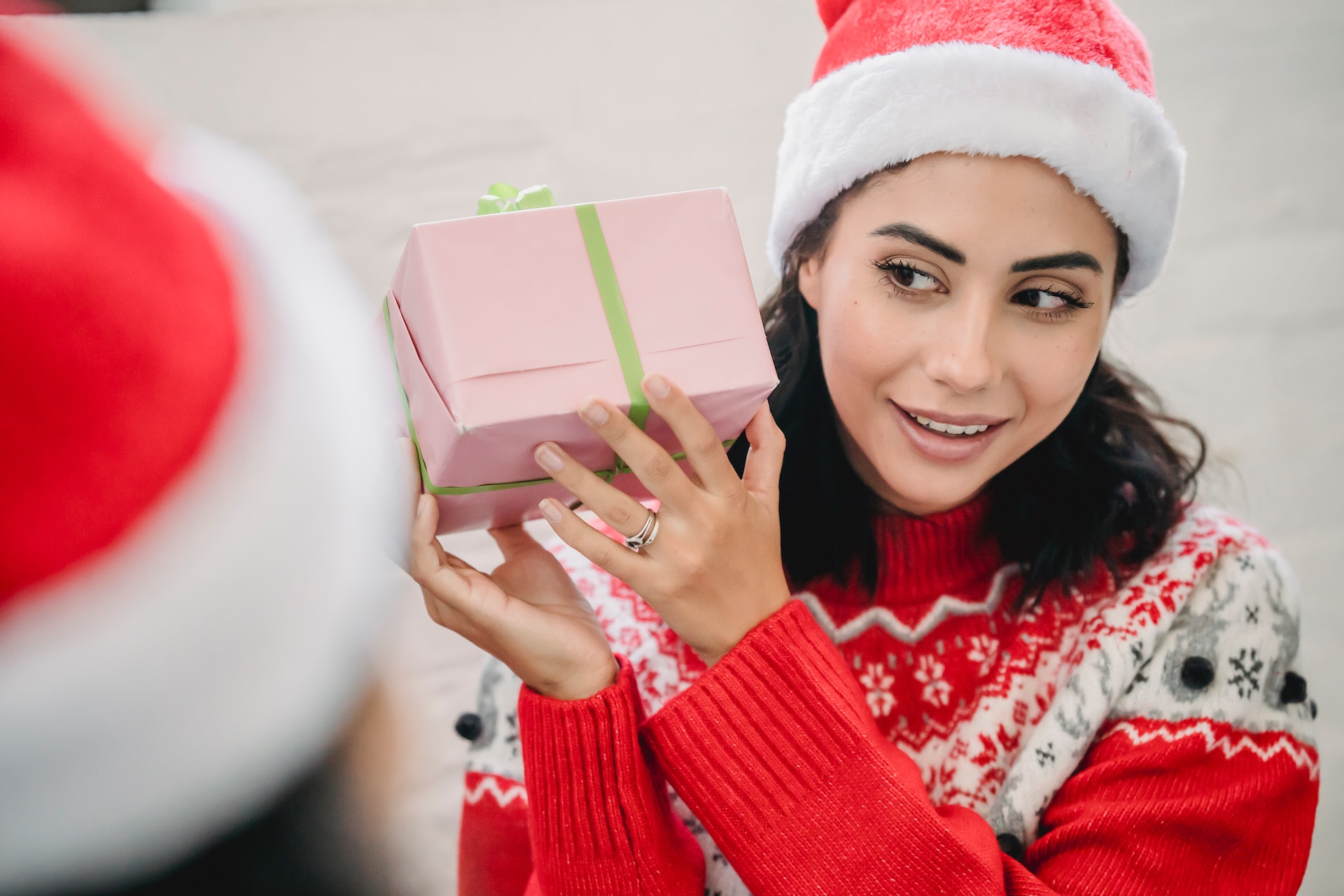 The image features a person wearing a red Santa hat and a red sweater with white snowflake patterns, suggesting a holiday theme, likely Christmas. The person is smiling and holding a pink gift wrapped with a green ribbon, seemingly looking at another person who is only partially visible and also appears to be wearing a red Santa hat. The partial view of the second person suggests that the photo might be taken from their perspective. The background is neutral and blurred, keeping the focus on the two people and the gift exchange moment.