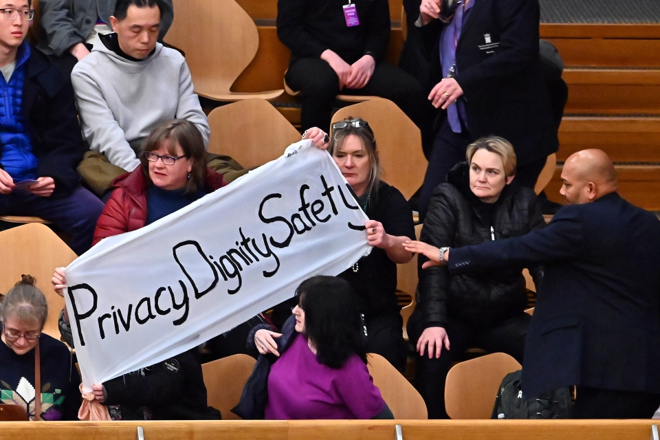 In the image, there is a group of people sitting in what appears to be an auditorium or a public meeting space. Several individuals are holding up a banner that reads \"Privacy Dignity Safety\". The action suggests that they are part of a demonstration or advocacy moment regarding these principles. One person, possibly a security guard or official, is reaching out towards the banner, possibly indicating an attempt to intervene or communicate with the banner holders. The rest of the audience is seated and appears to be watching the situation unfold. The context is not fully clear but it seems to be a public event where this message is being conveyed.