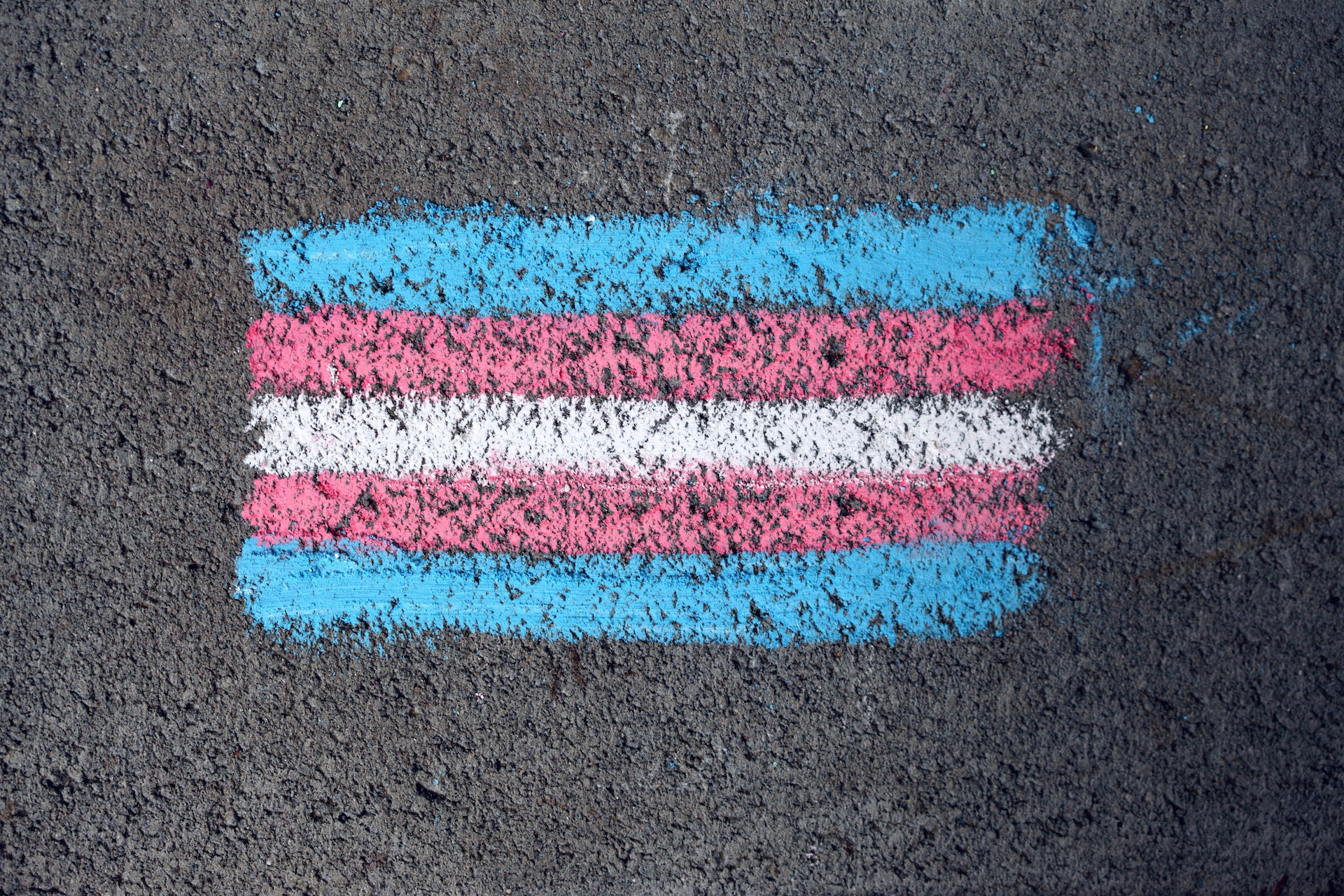 The image shows a horizontal stripe pattern painted on a rough surface, which appears to be asphalt due to its texture. The pattern consists of several colored horizontal bands: two light blue stripes on the top and bottom with a combination of pink, white, and another pink colored stripe in between, creating a five-striped block of color.\n\nThis pattern is distinctive and reminiscent of the Transgender Pride Flag, which represents the transgender community and includes these specific colors: blue (representing boys/men), pink (representing girls/women), and white (for those who are non-binary, transitioning or consider themselves having a neutral or undefined gender).