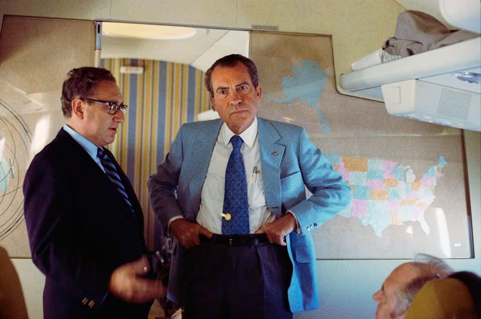 The image shows two men standing inside what appears to be an airplane cabin. The man on the right is wearing a light blue suit and is adjusting his belt, looking directly into the camera with a serious expression. The man on the left is wearing a dark suit and glasses and appears to be in conversation with the other man, or possibly just looking at him.\n\nIn the background, there's a map of the United States, suggesting this may be an office area or meeting space within the plane designed for work or discussions. Both men are dressed in professional attire, which might indicate they are business travelers, government officials, or individuals of a professional nature.\n\nThe image has a somewhat vintage feel to it, which might suggest it's from a few decades ago. It captures a candid moment during apparent travel, embodying a sense of motion and discussion.
