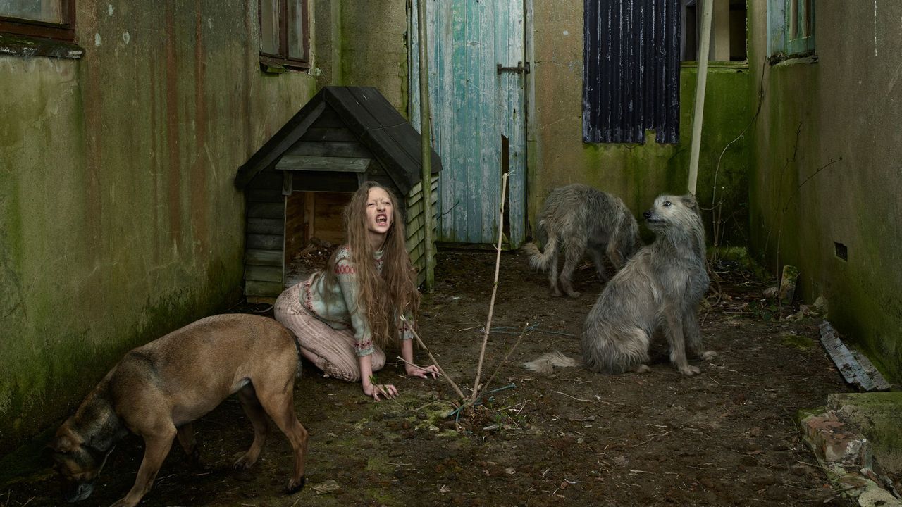 A woman sitting on the ground with two dogs, in front of a wooden house with a door and wall in the background. Sticks and leaves are scattered on the ground.